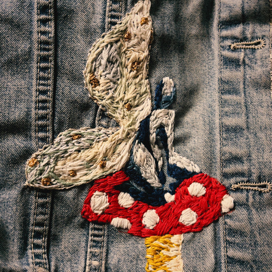 Close-up of a denim jacket featuring a detailed embroidered fairy and red mushroom motif on the front.