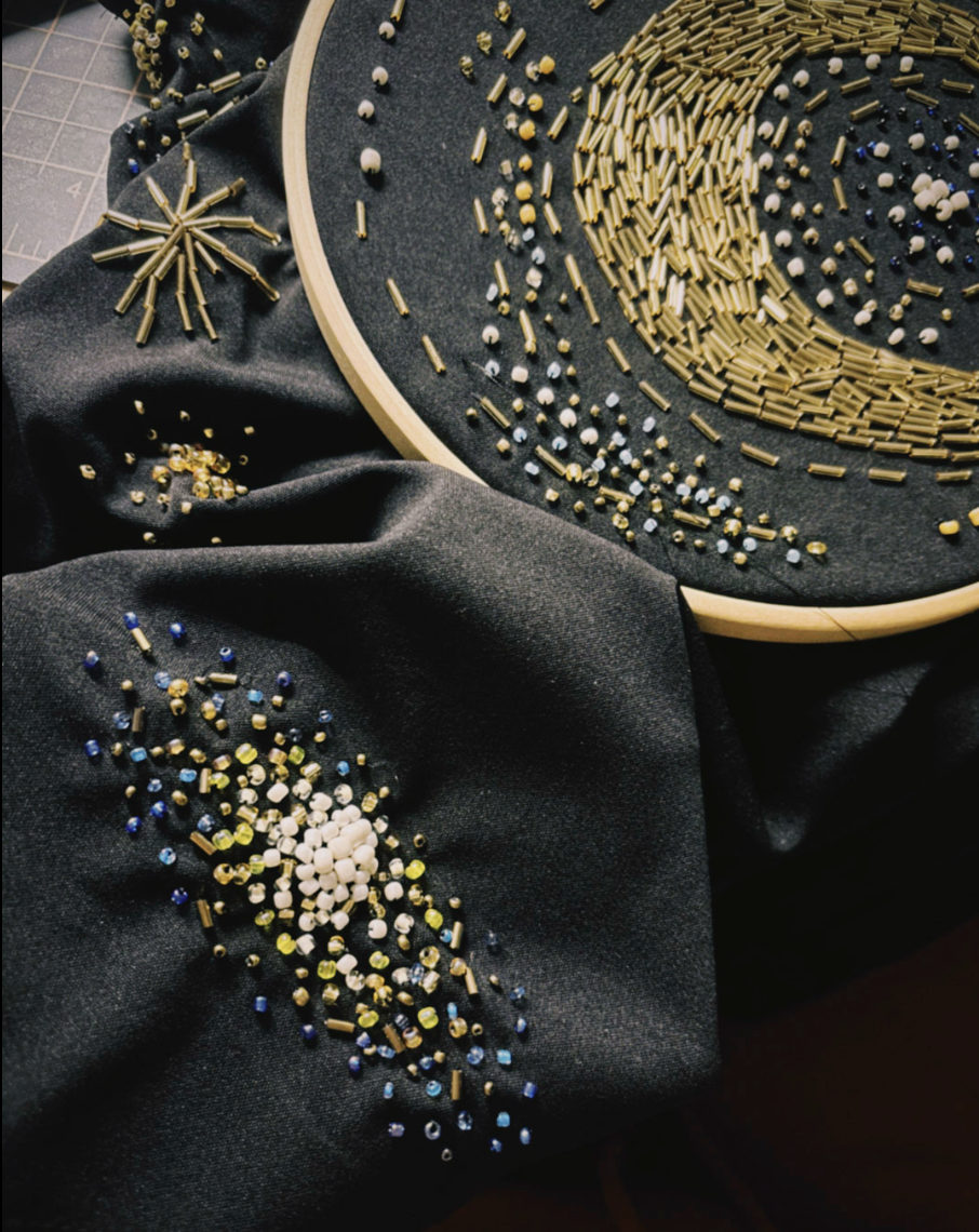 Close-up of a dark fabric surface decorated with embroidered crescent moon and star-like patterns made from metallic gold, blue, and white beads, with a wooden embroidery hoop holding matching thread and beads.