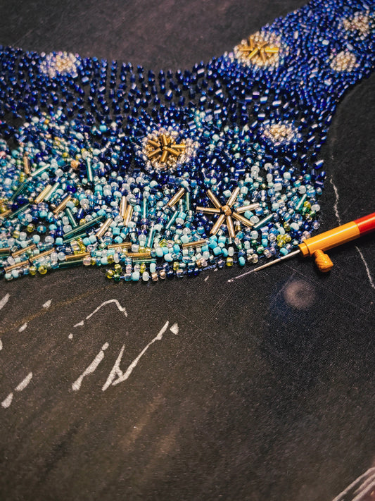 A close-up of a beaded textile or garment section featuring a blue and teal starry night pattern with gold accents, placed on a dark textured surface with a needle tool visible.