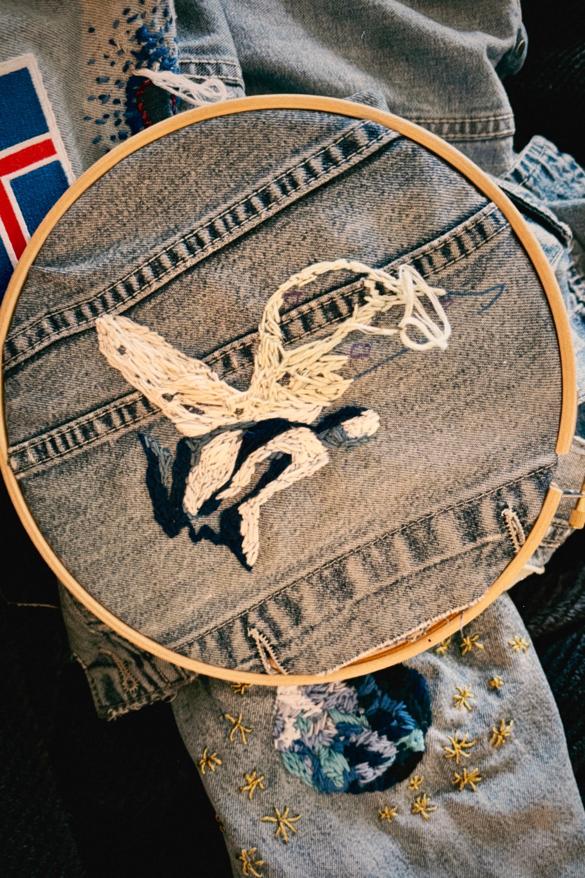 A close-up of a circular embroidery hoop holding a piece of light gray denim fabric with a white and black embroidered design, layered over other denim garments with floral patches and gold embroidery.