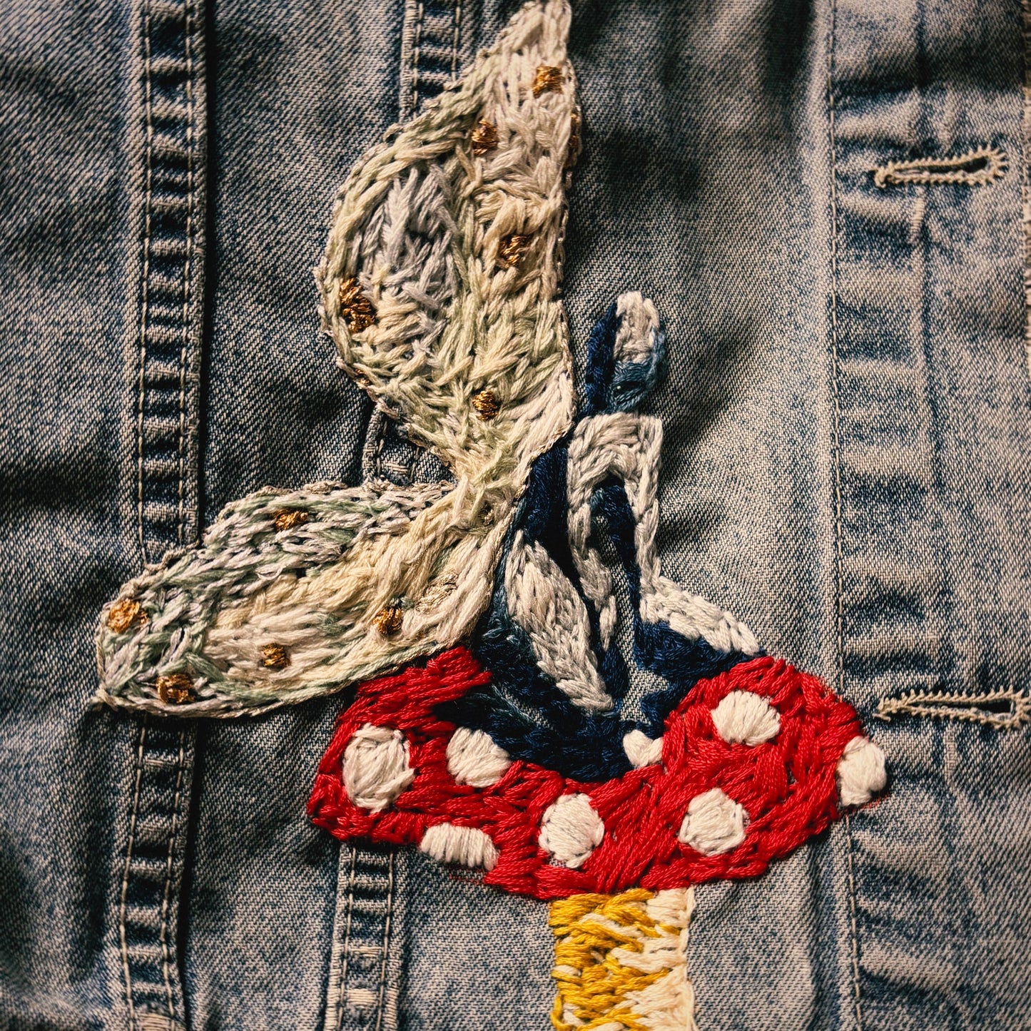 Close-up of a denim jacket featuring a detailed embroidered fairy and red mushroom motif on the front.