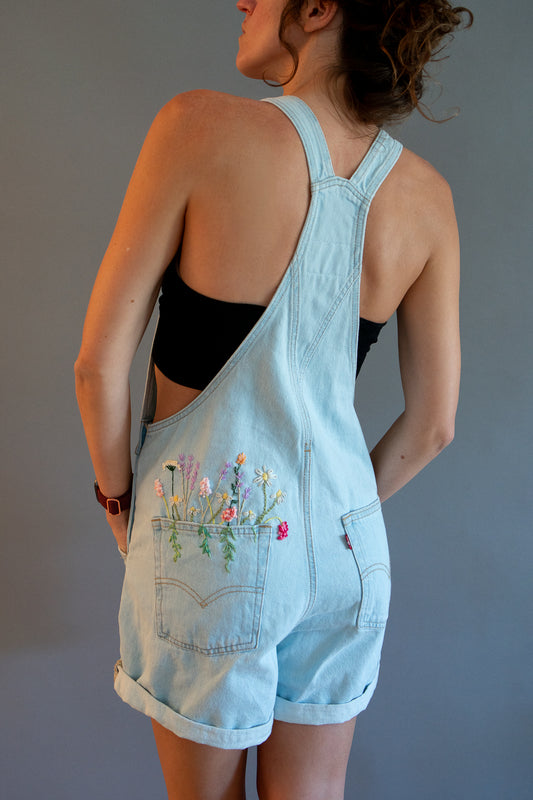 A woman shown from the back wearing light blue denim overall shorts with a floral embroidery on the pocket, styled over a black bandeau top against a plain gray background.
