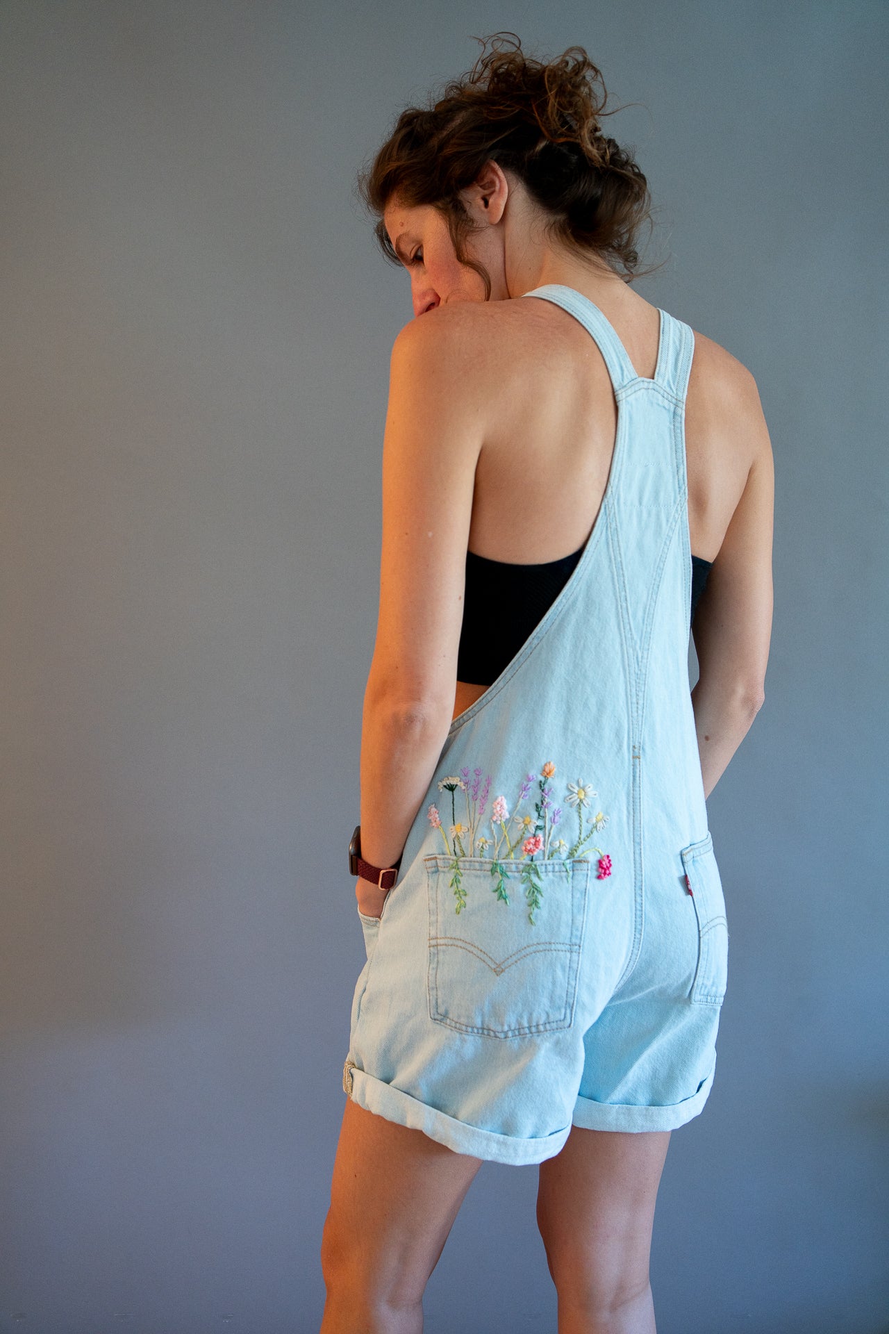 A woman wearing a light blue denim overall romper with floral embroidery on the back pocket, shown from behind against a plain gray studio background.
